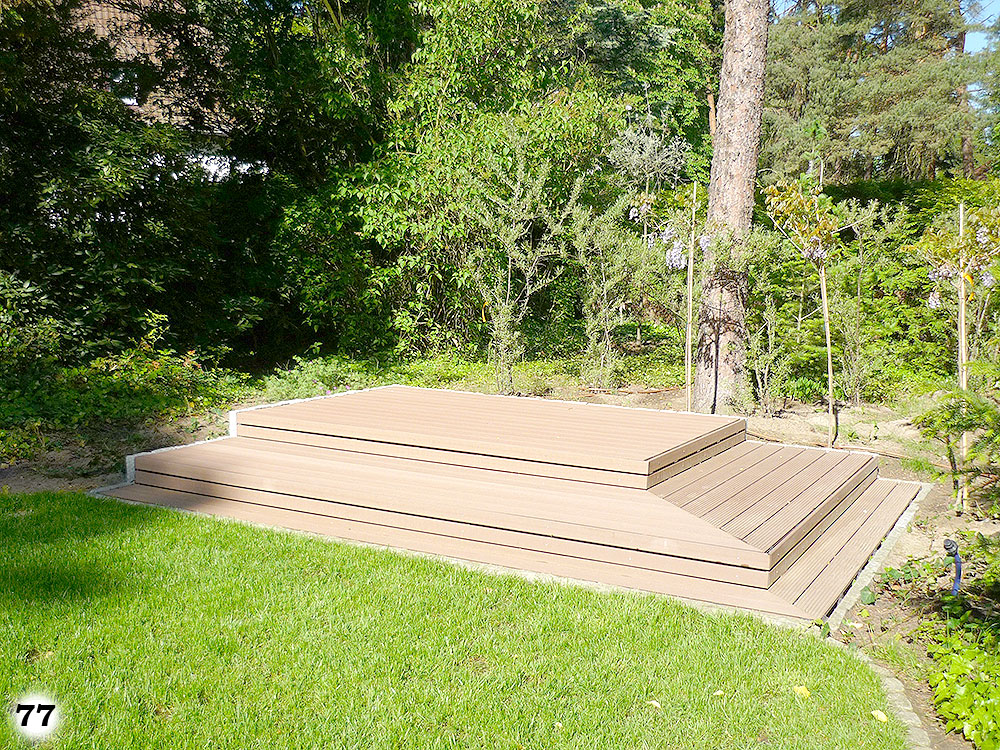 Ein einfacher Holzboden mit Stufen an einer Grünfläche die rundherum mit Bäumen versehen ist