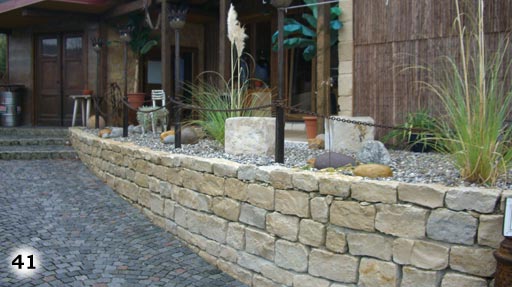 Mauer um einer Erhöhung auf der sich Steine, Pflanzen und ein Haus befindet
