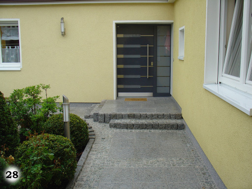 Weg aus quadratischen Steinplatten und kleinen, grauen, quadratischen Pflastersteinen führt zu einer Haustür mit zwei Stufen, welche aus kleinen, grauen, quadratischen Pflastersteinen sind