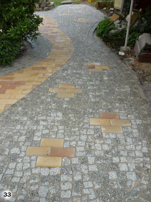 Weg aus kleinen, grauen, quadratischen Pflastersteinen mit einem Streifen und Mustern aus rötlichen und beachen Pflastersteinen