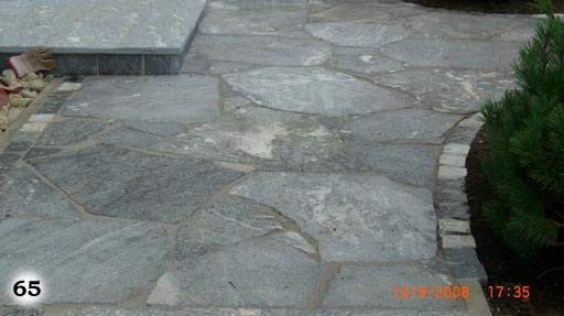 schmaler Weg aus hellen Steinplatten, umsäumt von kleinen, schwarzen, quadratischen Pflastersteinen, zwischen einer Rasenfläche und einem Beet